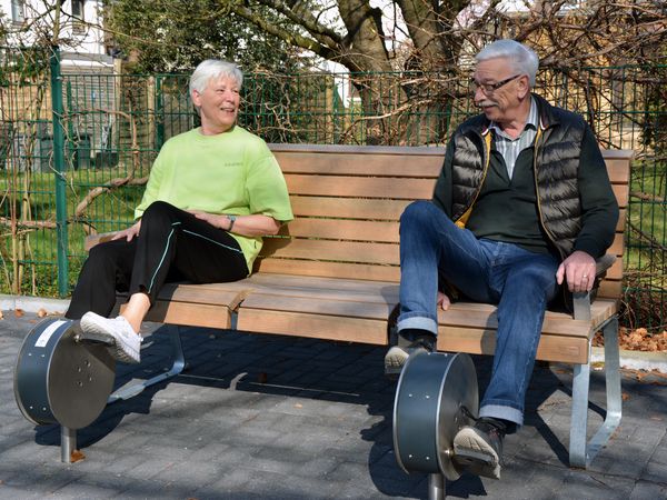 Ein älteres Ehepaar nutzt auf einer Bank sitzend spezielle Beintrainingsgeräte.